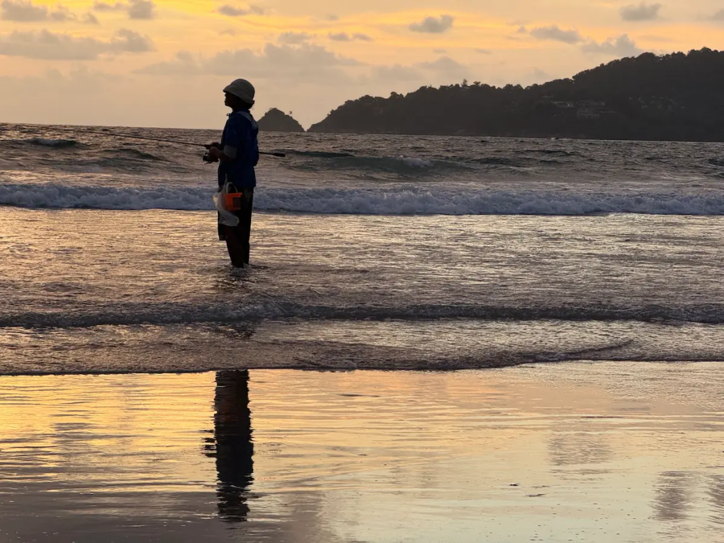 Travel photography tips illustrated by a fisherman standing in the surf at Patong Beach during sunset, showing how everyday moments tell a destination's story.